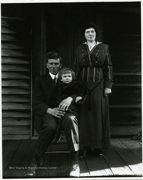 A father is holding his child and his wife is standing next to him on the front porch of their home in Helvetia, West Virginia.