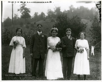Two young gentlemen standing outside between three ladies who are holding flowers. 