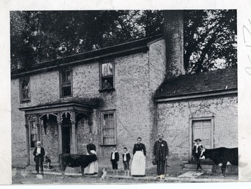 Two women and two gentlemen are standing in front of a farmhouse along with three children and two cattle in Huntington, West Virginia.