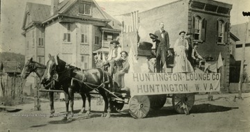 Men standing on a horse drawn float for Huntington Lounge Co. in Huntington, W. Va.  'Brick building at right is old Huntington Bank which was robbed by the James brothers gang in the 70's.  It later became the Huntington National Bank which merged with the First National Bank.  The tall man is Ham Dickey, once mayor of the city.'