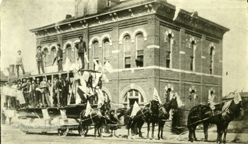 Men waving American flags while riding on a horse drawn cart during a Fourth of July parade.  They are passing 'Old City Hall. Scene of a hot election day pistol battle in the old days when men were men.'  