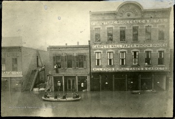 Men in a canoe float in front of the W. H. H. Holswade building. 'South Side of 3rd Ave. just West of 10th St.
