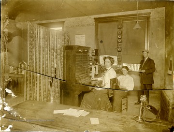 "Lewisburg Telephone Force" includes woman telephone operator, wearing a head set while sitting at the switchboard and two other unidentified persons. Note the column of "ringers" or bells attached to the middle window frame.