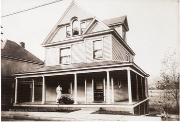 'Probably Irene Bucher Gorby on the porch.  This home was sold to Wilbur Jacob McColloch and Mildred Gorby McColloch in 1929.  The house to the left was owned by J. W. Postlethwaite.  It was later raised by 8-10 feet to be out of floods which plagued New Martinsville in the early 20th Century.  Flood waters never invaded the Poslethwaite living area, but reached to 8 feet in the living room of 714 Maple Ave., the window behind Mrs. Gorby.  This photo is about 1908 when the Gorby's were married.'