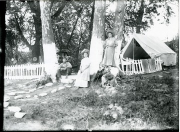 Two women and two boys are beside a white tent and picket fence.