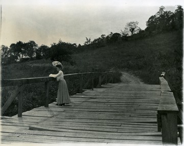 Woman standing on bridge spanning Falling Run. Pathway apparent on other side of bridge. Field leading to edge of trees.  