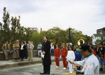 Speaker addressing public on New South Park bridge. Parade members can be seen behind speaker. Man holding microphone in lower right hand corner of picture. 
