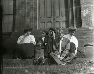 A group of young people sit on the steps in front of an brick building. 
