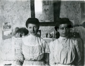 Two young ladies pose for a photo in what appears to be a room in a home. 