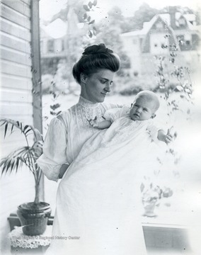 A woman stands holding a baby on an outside porch. Behind the woman, plants can be seen. 