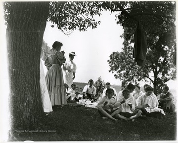 Men, women, and children seated on the ground eating a picnic lunch.