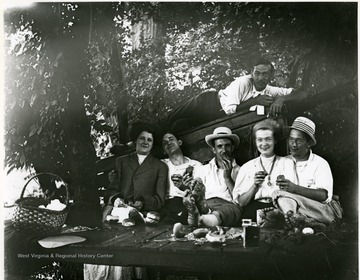 Three men and two women are sitting on a bench eating a picnic while a man is laying on top of the bench directly above them in Morgantown, West Virginia.