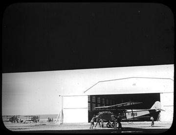 World War I Lantern Slide Show. Slide No. 22 in group of originally numbered slides.  American airfield with hanger and three planes.  A group of men are moving a biplane.  (negative no. 22-10976 is inscribed on slide)