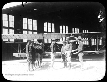 World War I Lantern Slide Show. Slide No. 15 in group of originally numbered slides.  American pilots standing at attention in front of biplane in hanger.  Frame is labelled with text saying 'Visual Bureau, University of Pittsburgh.'  (negative no. 28-10974 is inscribed on slide)<br />