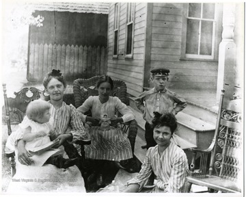 Three girls, a boy, and a baby seated outside.  The girls are sitting in ornate wooden and wicker chairs.