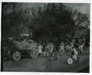 Children are riding their bicycles behind a decorated wagon float.