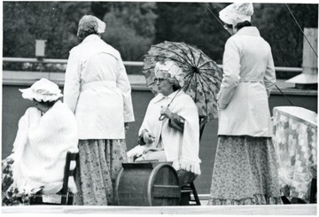 A group of older women in period clothes sits on chairs outside.
