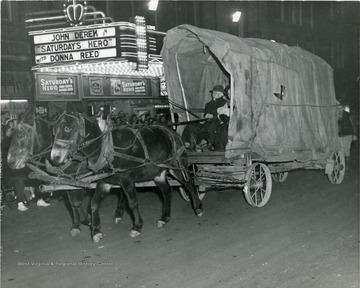 Covered wagon participating in a parade. Theater marquee advertising the movie, 'Saturday's Hero' is visible.