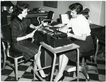 Two women sit at a desk with a typewriter.