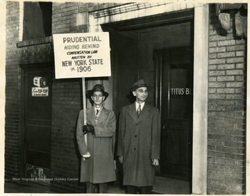 Two men stand in the doorway of the Titus Building.  One is holding a sign that reads 'Prudential Hiding Behind Compensation Law Written By New York State in 1906.'