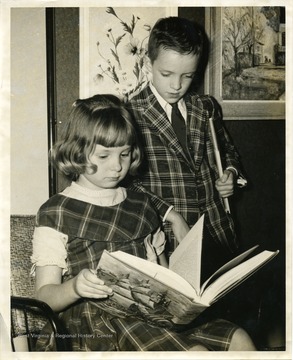A young girl is reading a thick book about ships and the sea while a young boy looks over her shoulder and glances at the book. 