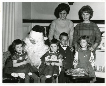 Three girls and a boy are being entertained by Santa Claus. Standing in the rear are Gail Nesius and Susan Feathers.