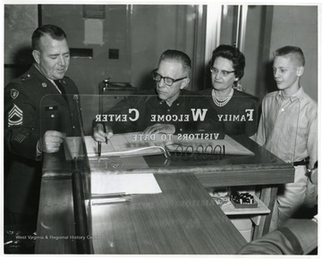 'Mr. L. B. Kerns from Morgantown, W. Va. signs the register at the Family Welcome Center in Fort Knox's Training Center after he had been informed that he and his family were the 100,000th visitors at the Center. Watching Mr. Kerns do the signing are his wife, and their son John. On the left is Master Sergeant Freddie Cox, the NonCommissioned Officer-in-Charge of the Center. The Kerns arrived November 5 to attend the graduation ceremony of their other son, James, who is completing his eight weeks of Basic Combat Training at Fort Knox with Company B, 13th Battalion, 4th Training Brigade. Their son will graduate November 6.''US Army Photograph'.
