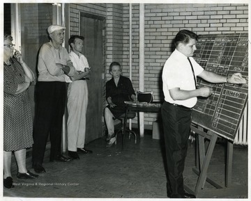 Dorthy Hiner, seated; Rex Everly, white shirt, standing; and Harold Wildman, wearing hat, standing; watch as votes are recorded on a chalk board.