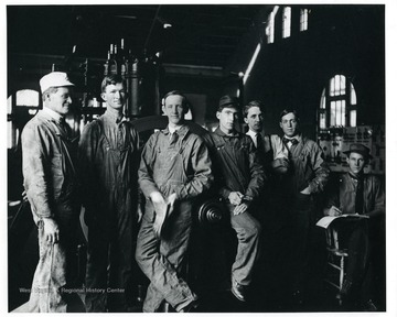 A group of seven men in work clothes inside an unidentified factory.