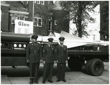 Amry men standing in front of a missle in their uniforms. 
