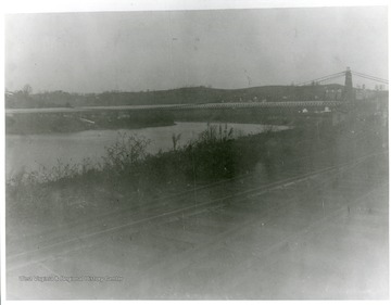 'A view of Morgantown Suspension Bridge. The Baltimore and Ohio Railroad tracks are in the foreground. West Virginia University buildings are at the top right.'