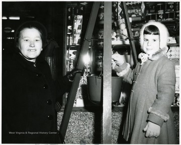 A women and a little girl standing next to a Salvation Army Christmas kettle in front of a store.