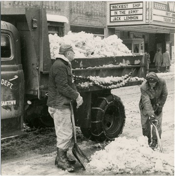 Two men shoveling snow into the back of a truck. You can see the Warner movie theatre in the background showing 'Wackiest Ship in the Army' starring Jack Lemmon. 