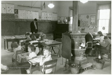 Interior view of one room elementary school with coal burning stove lit and children of different ages.