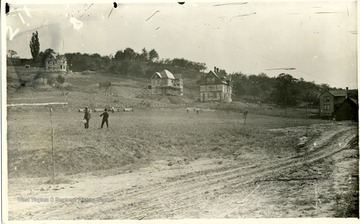 Two unidentified men stand in the middle of the athletic field near Fraternity Row on North High Street in Morgantown, West Virginia. This photograph was taken when the first houses were being built along Fraternity Row.