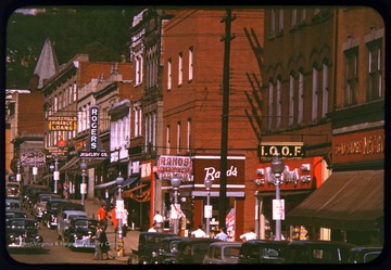 A view of High Street, looking north, in downtown Morgantown.  This view features automobiles and pedestrians during a busy time of day.  Shop signs are clearly in view, including Rands Drugs, Rogers Jewelry Company, and Comuntzis Restaurant.