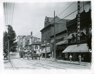 A horse and buggy and a streetcar are driving down High Street near Hirschmans Store. They are near the intersection of Walnut and High Street in Morgantown, West Virginia.