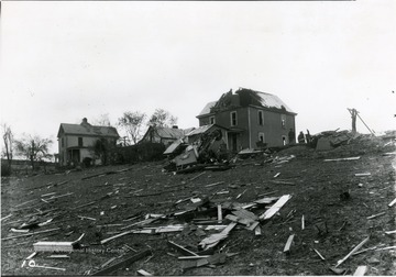 An area damaged by a windstorm. The roof of the house in the center has been torn apart, and in the front, a car is flipped upside down.