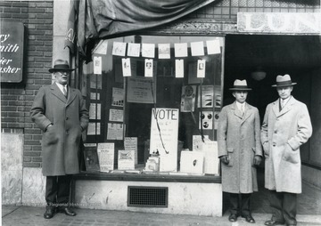 Three men standing outside of a window display urging people to vote.