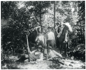 Group portrait of men in the woods.  Two have guns, one has an axe, another is carrying water and a coffee pot, two are on horse back and another is posed on a felled tree.