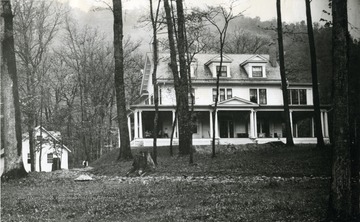 A view of the John Raine home in Rainelle. People on a porch swing converse while  someone works in the yard to the left of the home.
