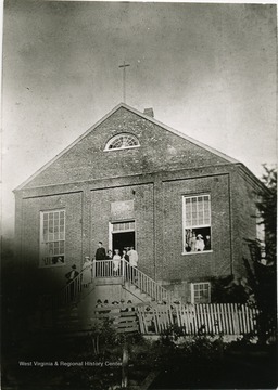 A view of the Catholic Church in Weston where school was conducted in its basement. People are gathered on the steps and in front of the church, and looking out the window. The church is located on East 2nd Street and Court Street. 'It is now the Columbian Club.'