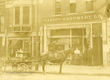 A view of the Marion Hardware Company building on Main Street in Fairmont. A horse-drawn carriage in front of the store.  'Standing in the doorway are Miss Olive Brand, H. J. Jones, and L. C. Boice.' 