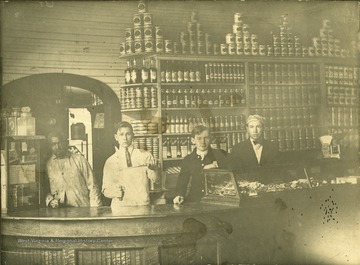 Olie, second from left, stands behind a counter sharpening a knife at Matoaka. An African American man stands to his right, and to his left are two other young men who also work at the store.