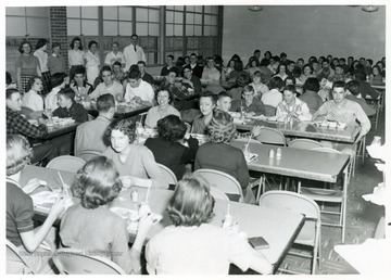 "Alumns serving Rampfeast, 1956, to buy cafeteria equipment.  Sale was a success.  Standing by the wall, left to right, are girls who served food.  Barbara Cogar, Connie Ware, Frances Gladwell, Maxine House.  Cooks who prepared food are Lafa Hosey, Mrs. Olive Nichols, and Robert G. Andrick who was manager of the cafeteria from 1955 to 1960.  At the center of the room is Miss Aretta Summers and Mrs. Ruth Morton Himes, both reflecting appreciation for the new high school building.  They were teachers of whom the school was proud."