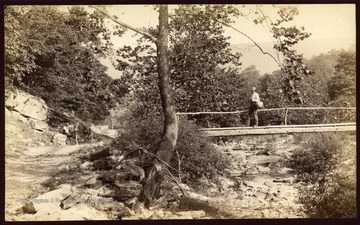 A fisherman is on the foot bridge and a man with surveying equipment is seated beside the road.