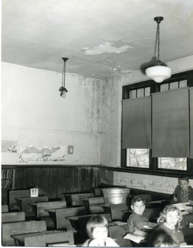 A large tub sits under a leaking ceiling. The children seated a safe distance from the damaged ceiling, focus on the camera. The class and school were not identified.