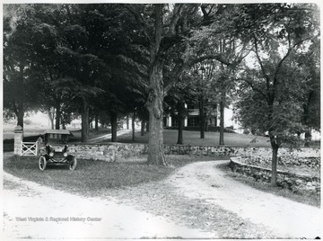 An early model automobile is parked under a large tree beside the driveway.