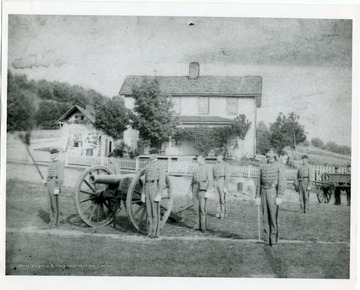 Military personnel are standing at attention in front of a cannon and Fife Cottage, West Virginia University.