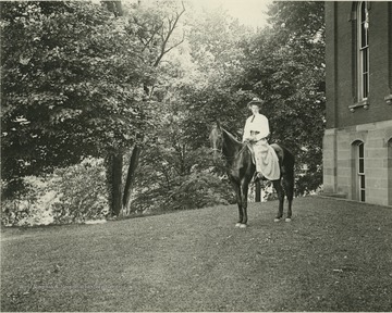 Leila Jesse Frazier, of Upper Norword in Surrey, England and an 1899 graduate of the WVU Law School, rides 'man fashion' or astride,  near Woodburn Hall. A contemporaneous newspaper account depicts Frazier’s journey to Morgantown to begin her law studies, indicating that she put her husband, James C. Frazier, on the train in Martinsburg, and set off unaccompanied on horseback across the mountains.  She arrived several days later,wearing a black riding habit with a divided skirt, riding ';man fashion', carrying a brace of revolvers, and 'armed with a most remarkable amount of courage and daring'. Frazier was president of the Woman’s League of WVU, the first women’s organization on campus. Information from Becky Lofstead, 'Trailblazers at the College of Law' in WVU Alumni Magazine, Winter 2000, p.18.